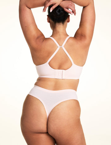 On a light studio backdrop, Jennifer wears the Cloud Cotton Highwaist Thong in Dogwood with a matching bra in Dogwood. The back view highlights the thong back coverage, smooth waistband, and convertible racerback straps on the bra.