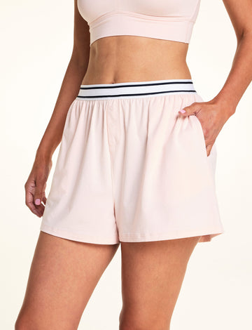 On a light studio backdrop, Meagan wears the Cloud Cotton Short in Dogwood with a matching Dogwood bralette. The close-up highlights the elastic logo waistband, functional snap fly detail, relaxed fit, and side pocket. sizing: Meagan wears size Medium