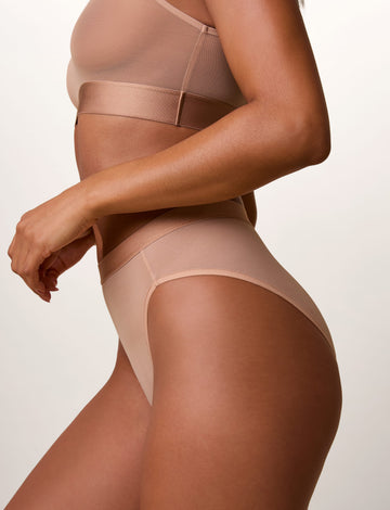 On a light studio backdrop, Ally wears the ComfortMesh™ Highwaist Brief in Taupe with the matching ComfortMesh™ Scoop Bralette. Shown from a side angle, the brief features sheer mesh fabric, a high-rise fit, and a smooth waistband.
