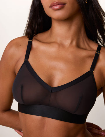 On a light studio backdrop, Ally wears the ComfortMesh™ Triangle Bralette in Black. A close-up view highlights the sheer mesh fabric, V-neckline, adjustable straps with gold-tone hardware, and wide comfort band.