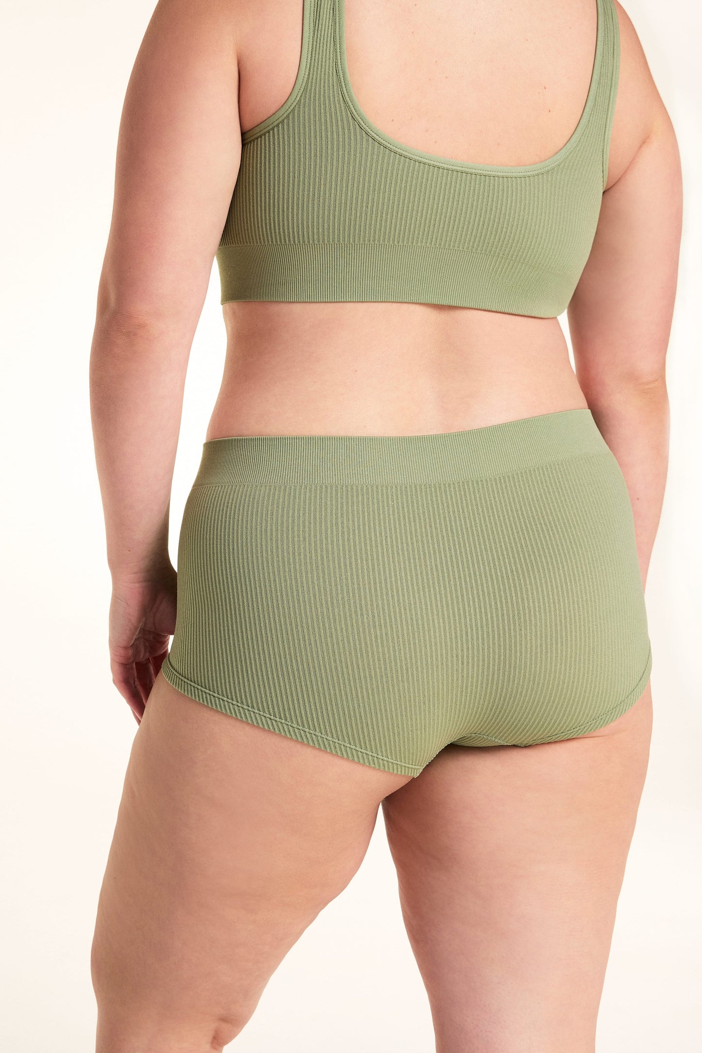 On a white studio backdrop, Alessandra models the back of the Lounge Rib Sleepshort in Dill with the matching Lounge Rib bralette, showing the ribbed fabric, seamless construction, and full back view.