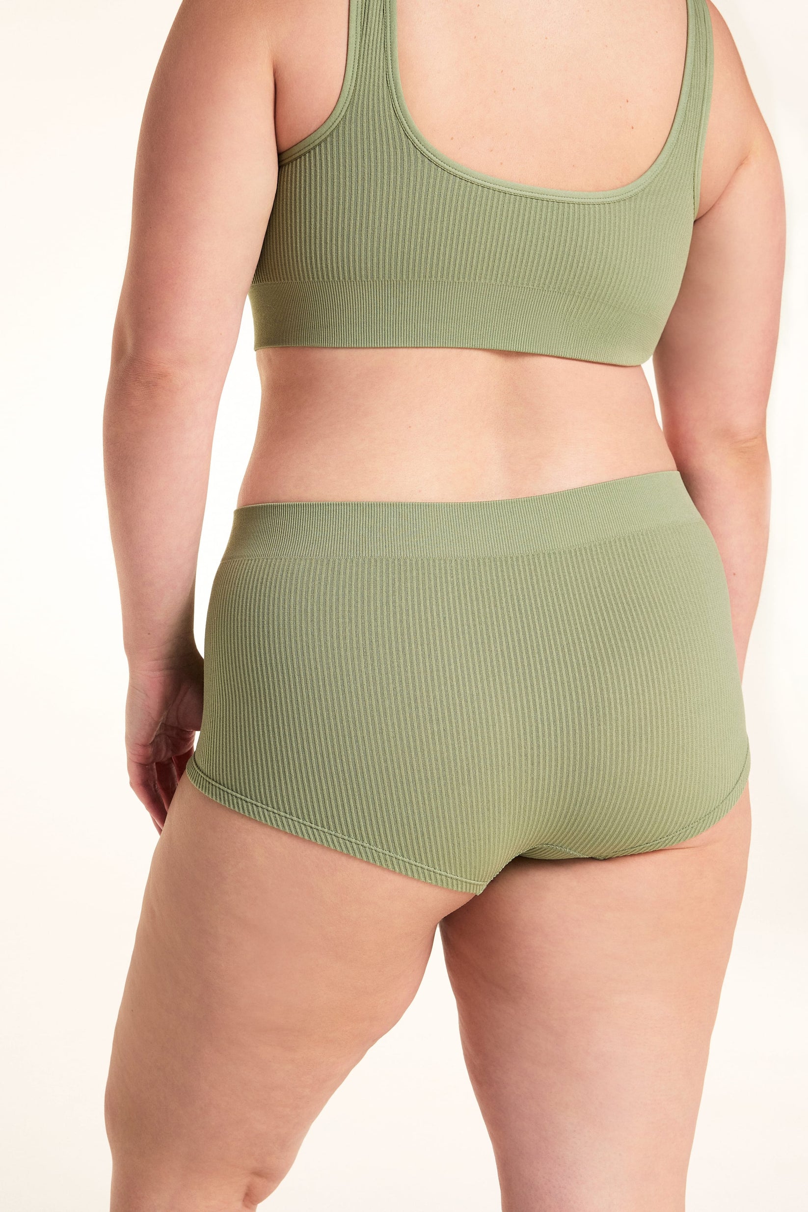 On a white studio backdrop, Alessandra models the back of the Lounge Rib Sleepshort in Dill with the matching Lounge Rib bralette, showing the ribbed fabric, seamless construction, and full back view.