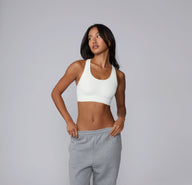 On a neutral studio backdrop, Amber stands wearing the Seamless Racerback Sports Bra in White, paired with gray sweatpants. The smooth scoop neckline, wireless support, and clean racerback silhouette are visible from the front.