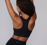 On a neutral studio backdrop, Courtney is shown from the back wearing the Seamless Racerback Sports Bra in Black with matching leggings. The racerback silhouette, breathable textured panels, and wide wireless band with a subtle Harper Wilde logo are visible.