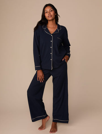 On a neutral studio backdrop, Victoria wears the Perfect Pima Pajama Long Sleeve Shirt in Navy with matching pajama pants. The full-body view showcases the relaxed silhouette, button-front design, chest pocket, and contrast piping details throughout the set.