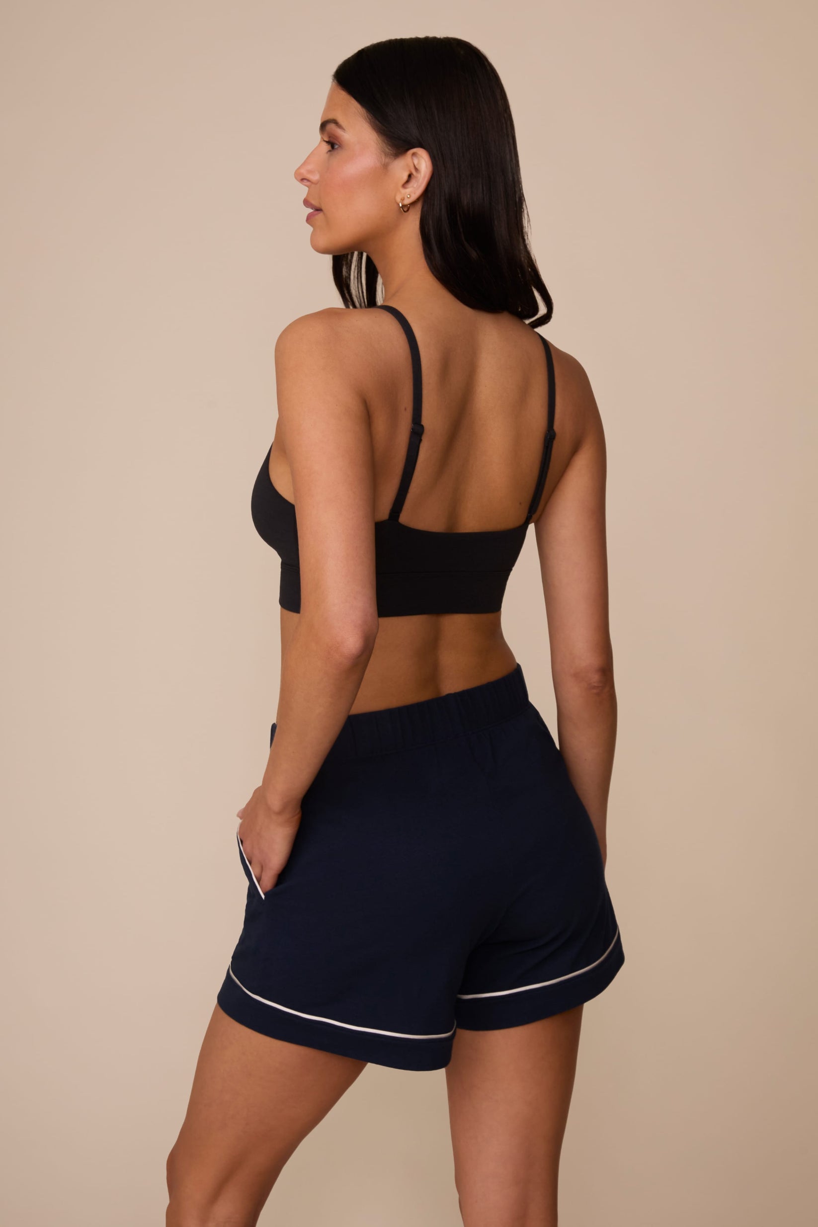 On a neutral studio backdrop, Marcela wears the Perfect Pima Pajama Short in Navy with a black bralette. The back view showcases the relaxed silhouette, elastic waistband, and contrast piping detail at the leg openings.