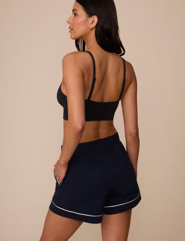 On a neutral studio backdrop, Marcela wears the Perfect Pima Pajama Short in Navy with a black bralette. The back view showcases the relaxed silhouette, elastic waistband, and contrast piping detail at the leg openings.