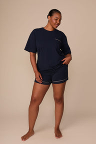On a warm beige studio backdrop, Zarria wears the Perfect Pima Pajama T-Shirt in a deep navy color with matching pajama shorts. The full-body view showcases the relaxed silhouette, crew neckline, short sleeves, and coordinating pajama set with contrast piping at the pocket and shorts hem.