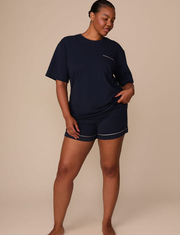 On a warm beige studio backdrop, Zarria wears the Perfect Pima Pajama T-Shirt in a deep navy color with matching pajama shorts. The full-body view showcases the relaxed silhouette, crew neckline, short sleeves, and coordinating pajama set with contrast piping at the pocket and shorts hem.