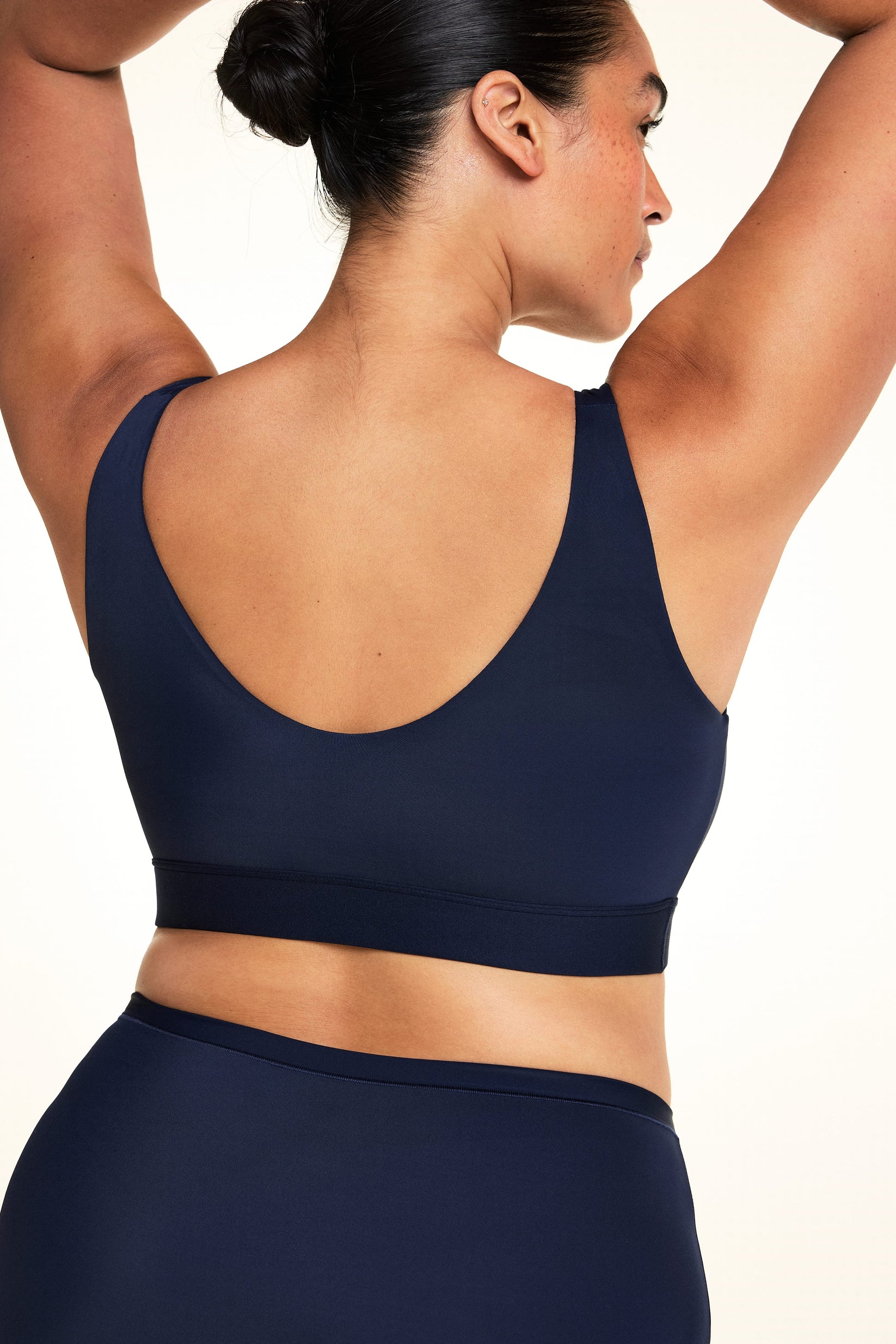 On a light studio backdrop, Jennifer is shown from the back wearing the Bliss Bralette in Navy with matching underwear. Her arms are raised overhead, highlighting the scoop back, wide band, and smooth microfiber fabric.