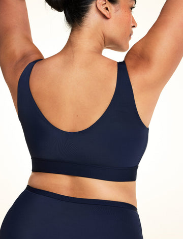 On a light studio backdrop, Jennifer is shown from the back wearing the Bliss Bralette in Navy with matching underwear. Her arms are raised overhead, highlighting the scoop back, wide band, and smooth microfiber fabric.