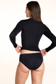 On a white studio backdrop, Laura wears the Bliss Long-Sleeve Tee and Bliss Low-Rise Bikini in Black, shown from the back while lifting the tee to show the bikini’s back coverage.