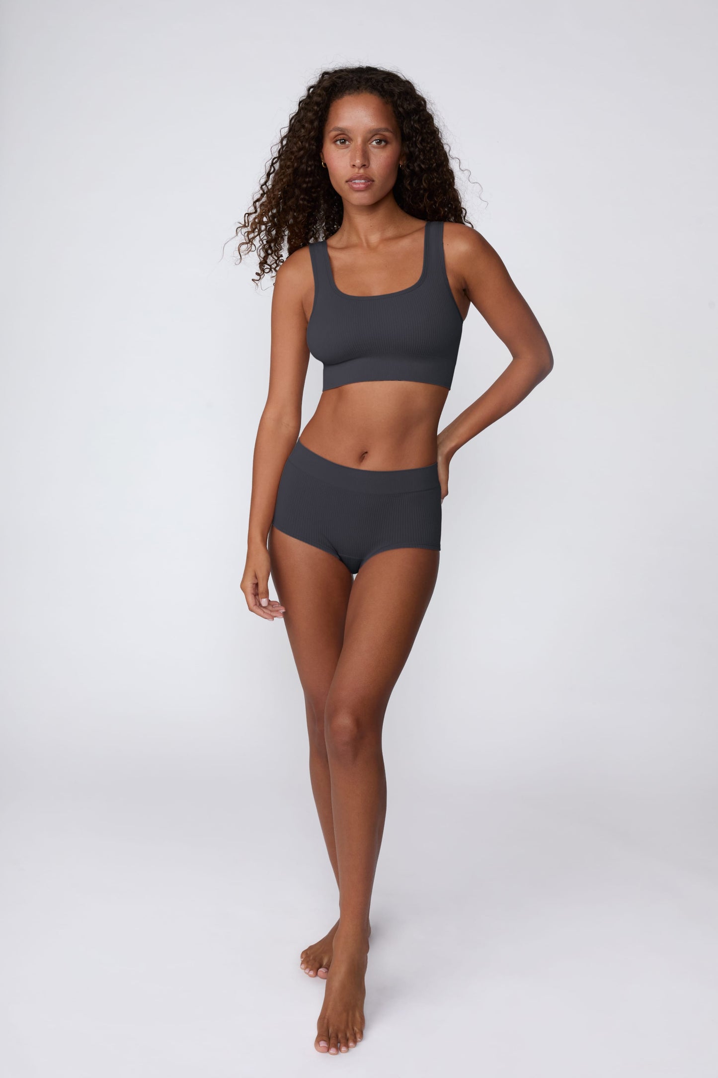 On a light gray studio backdrop, Courtney wears the Ribbed Seamless Boyshort in Charcoal with a matching ribbed bralette. A full-body view highlights the high-rise waistband, boyshort silhouette, and ribbed texture.