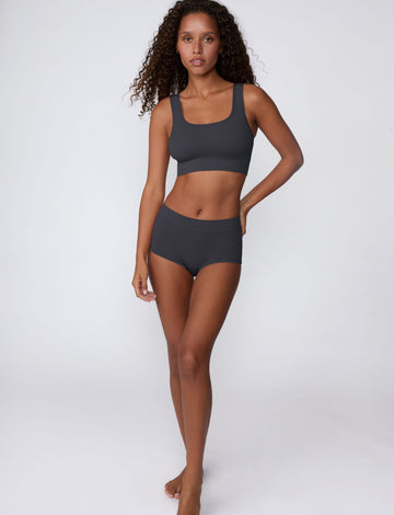 On a light gray studio backdrop, Courtney wears the Ribbed Seamless Boyshort in Charcoal with a matching ribbed bralette. A full-body view highlights the high-rise waistband, boyshort silhouette, and ribbed texture.
