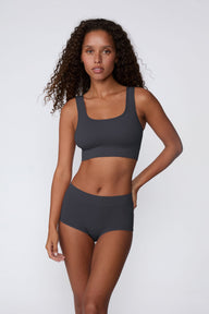 On a light gray studio backdrop, Courtney wears the Ribbed Seamless Scoop Bralette in Charcoal with a matching ribbed bottom. A full-body view highlights the scoop neckline, wide straps, ribbed texture, and supportive underband.