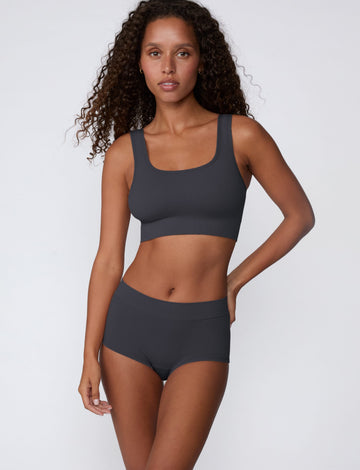 On a light gray studio backdrop, Courtney wears the Ribbed Seamless Scoop Bralette in Charcoal with a matching ribbed bottom. A full-body view highlights the scoop neckline, wide straps, ribbed texture, and supportive underband.