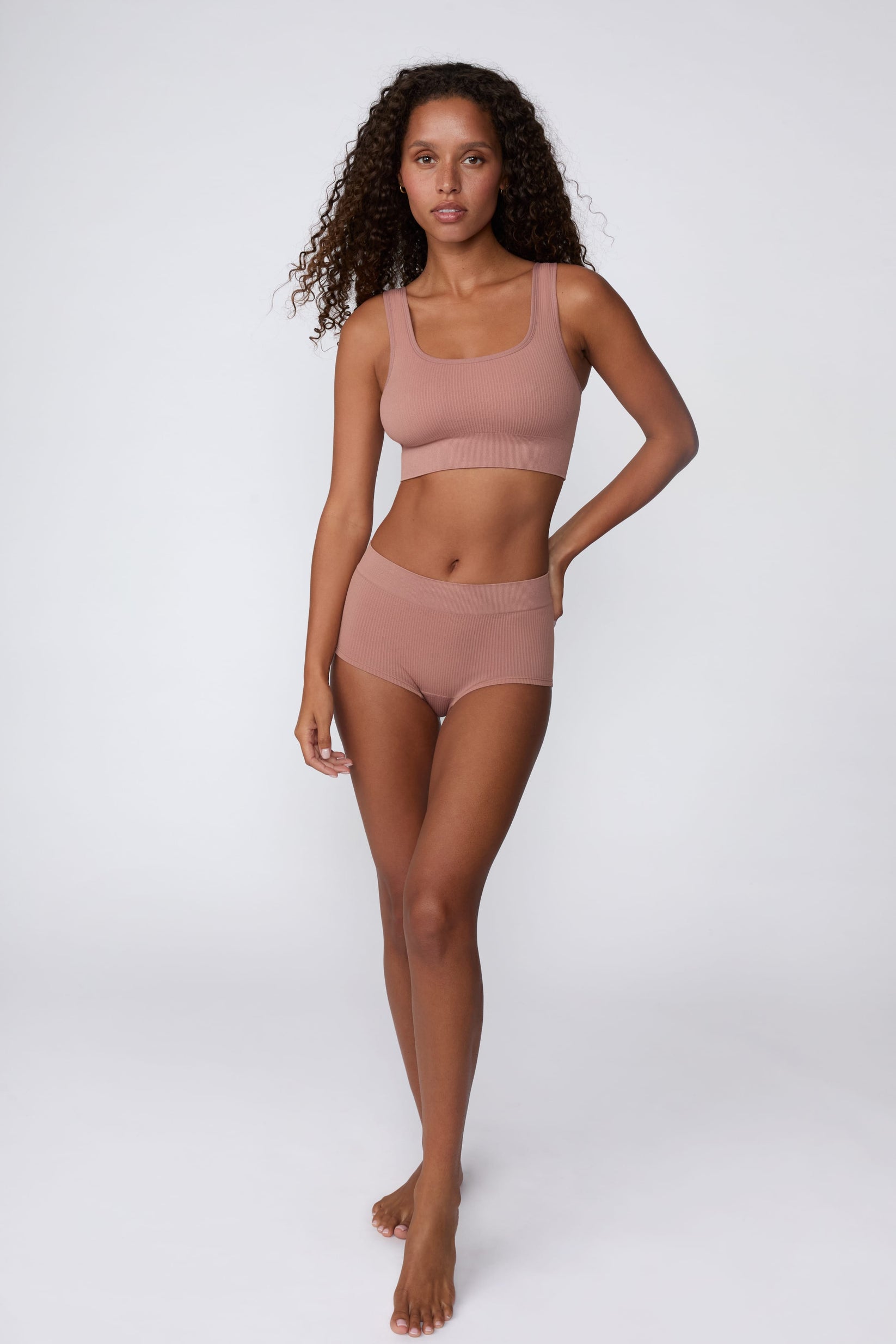 On a light gray studio backdrop, Courtney wears the Ribbed Seamless Scoop Bralette in Plum with a matching ribbed bottom. A full-body view highlights the scoop neckline, wide straps, ribbed texture, and supportive underband.