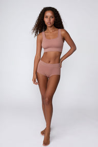 On a light gray studio backdrop, Courtney wears the Ribbed Seamless Scoop Bralette in Plum with a matching ribbed bottom. A full-body view highlights the scoop neckline, wide straps, ribbed texture, and supportive underband.