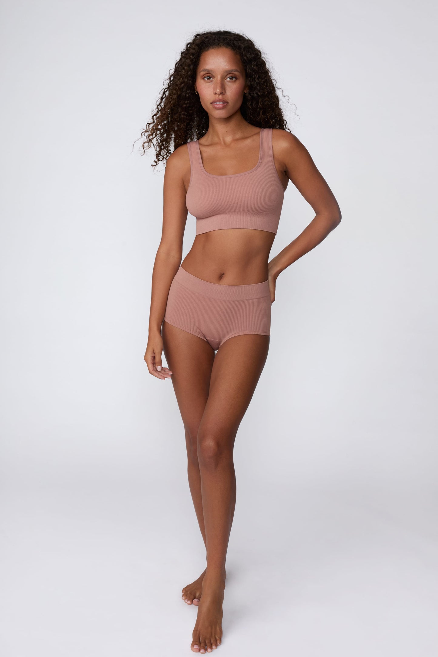On a light gray studio backdrop, Courtney wears the Ribbed Seamless Scoop Bralette in Plum with matching ribbed bottoms. A full-body view highlights the scoop neckline, wide straps, and ribbed texture.