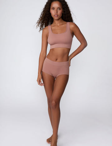 On a light gray studio backdrop, Courtney wears the Ribbed Seamless Scoop Bralette in Plum with matching ribbed bottoms. A full-body view highlights the scoop neckline, wide straps, and ribbed texture.