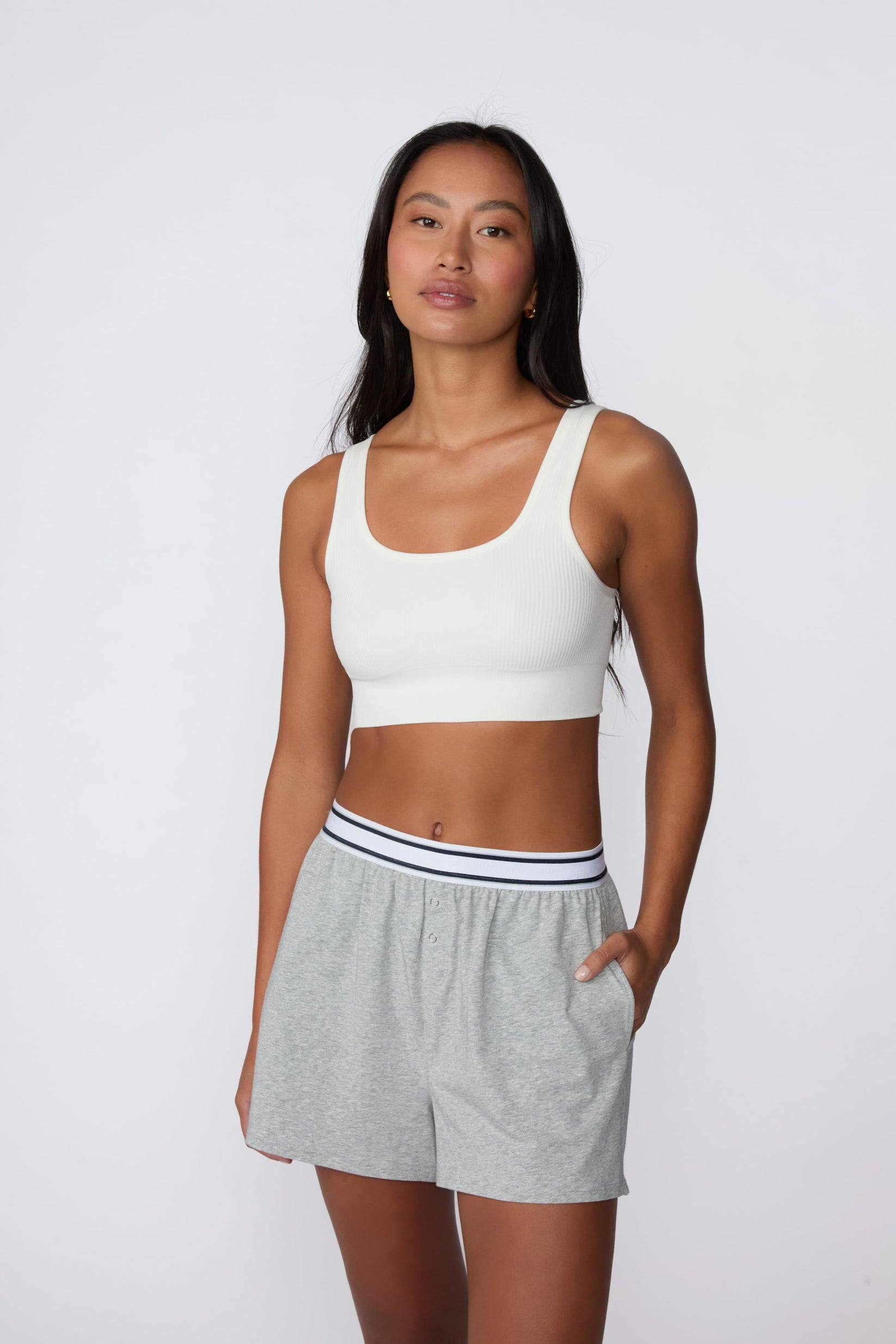 On a light gray studio backdrop, Amber wears the Ribbed Seamless Scoop Bralette in White with gray lounge shorts. A full-body view highlights the scoop neckline, wide straps, ribbed texture, and supportive underband.