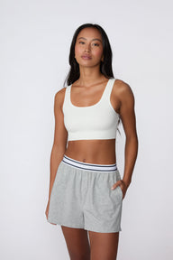 On a light gray studio backdrop, Amber wears the Ribbed Seamless Scoop Bralette in White with gray lounge shorts. A full-body view highlights the scoop neckline, wide straps, ribbed texture, and supportive underband.