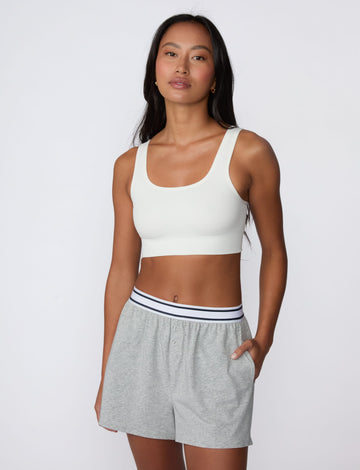On a light gray studio backdrop, Amber wears the Ribbed Seamless Scoop Bralette in White with gray lounge shorts. A full-body view highlights the scoop neckline, wide straps, ribbed texture, and supportive underband.