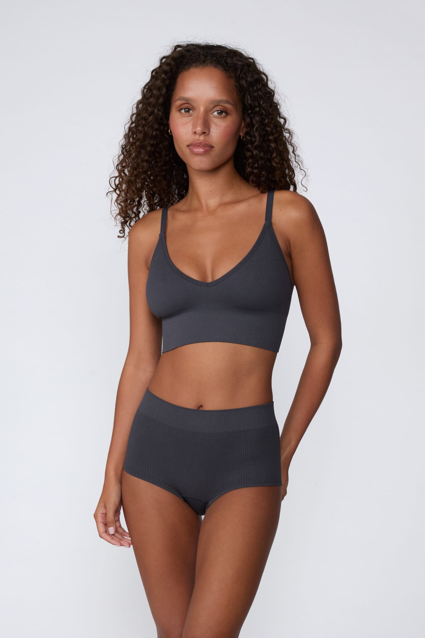 On a light gray studio backdrop, Courtney wears the Ribbed Seamless Triangle Bralette in Charcoal with a matching ribbed bottom. A full-body view highlights the ribbed fabric, V-neckline, adjustable straps, and wide supportive underband.