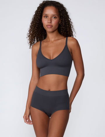 On a light gray studio backdrop, Courtney wears the Ribbed Seamless Triangle Bralette in Charcoal with a matching ribbed bottom. A full-body view highlights the ribbed fabric, V-neckline, adjustable straps, and wide supportive underband.