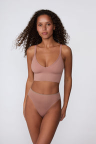 On a light gray studio backdrop, Courtney wears the Ribbed Seamless Triangle Bralette in Plum with matching underwear. The front view shows the deep V neckline, ribbed seamless construction, adjustable straps, and longline silhouette.