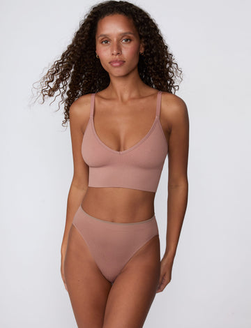 On a light gray studio backdrop, Courtney wears the Ribbed Seamless Triangle Bralette in Plum with matching underwear. The front view shows the deep V neckline, ribbed seamless construction, adjustable straps, and longline silhouette.