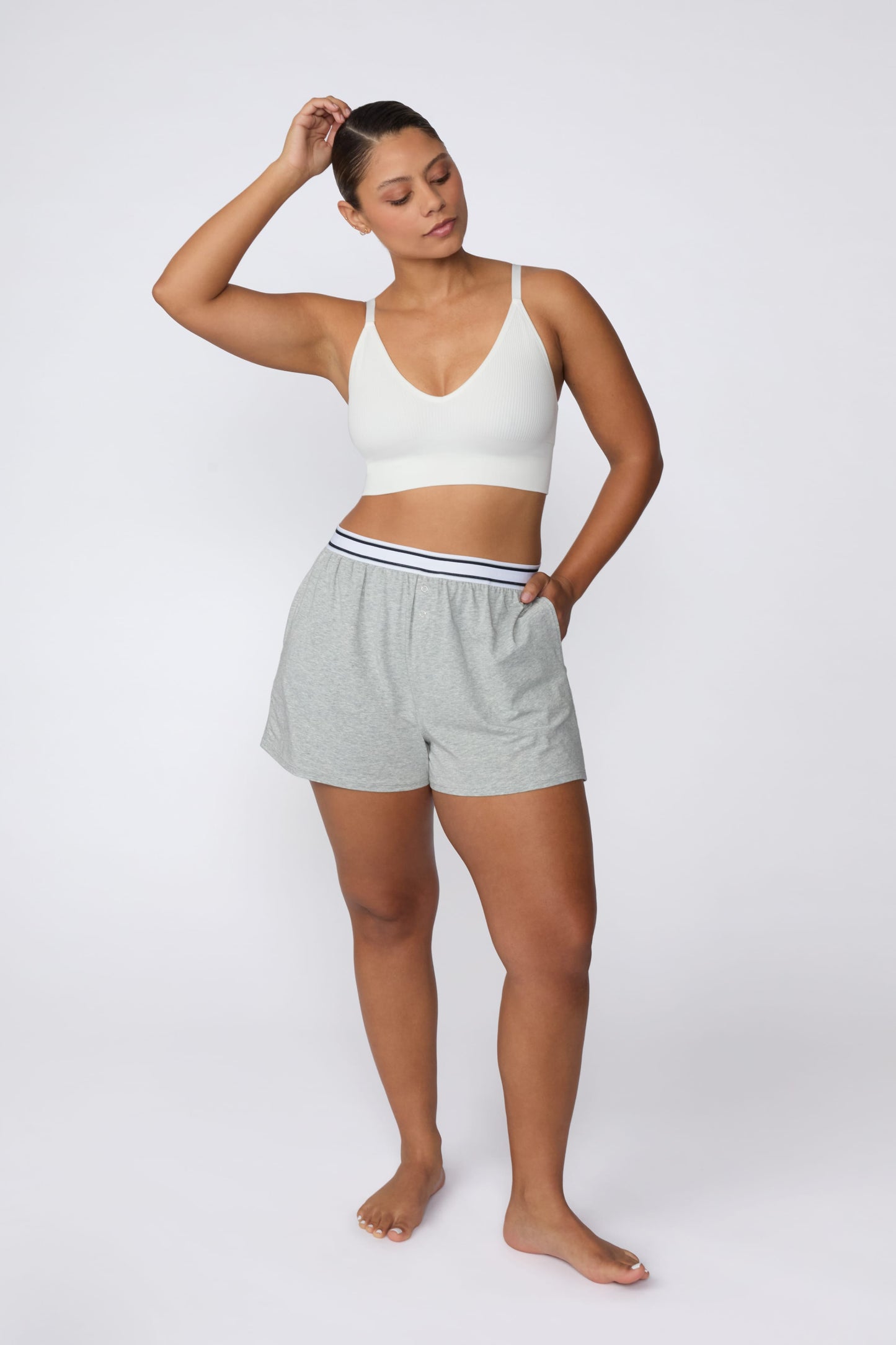 On a light gray studio backdrop, India wears the Ribbed Seamless Triangle Bralette in White with gray lounge shorts. A full-body view highlights the deep V-neckline, adjustable straps, ribbed texture, and wide supportive underband.