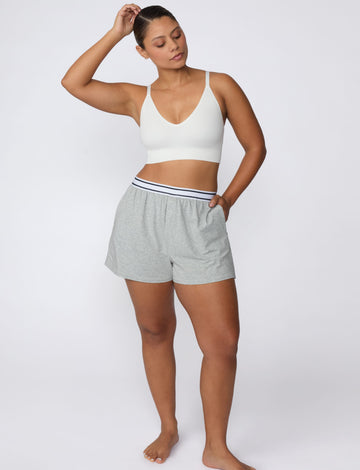 On a light gray studio backdrop, India wears the Ribbed Seamless Triangle Bralette in White with gray lounge shorts. A full-body view highlights the deep V-neckline, adjustable straps, ribbed texture, and wide supportive underband.