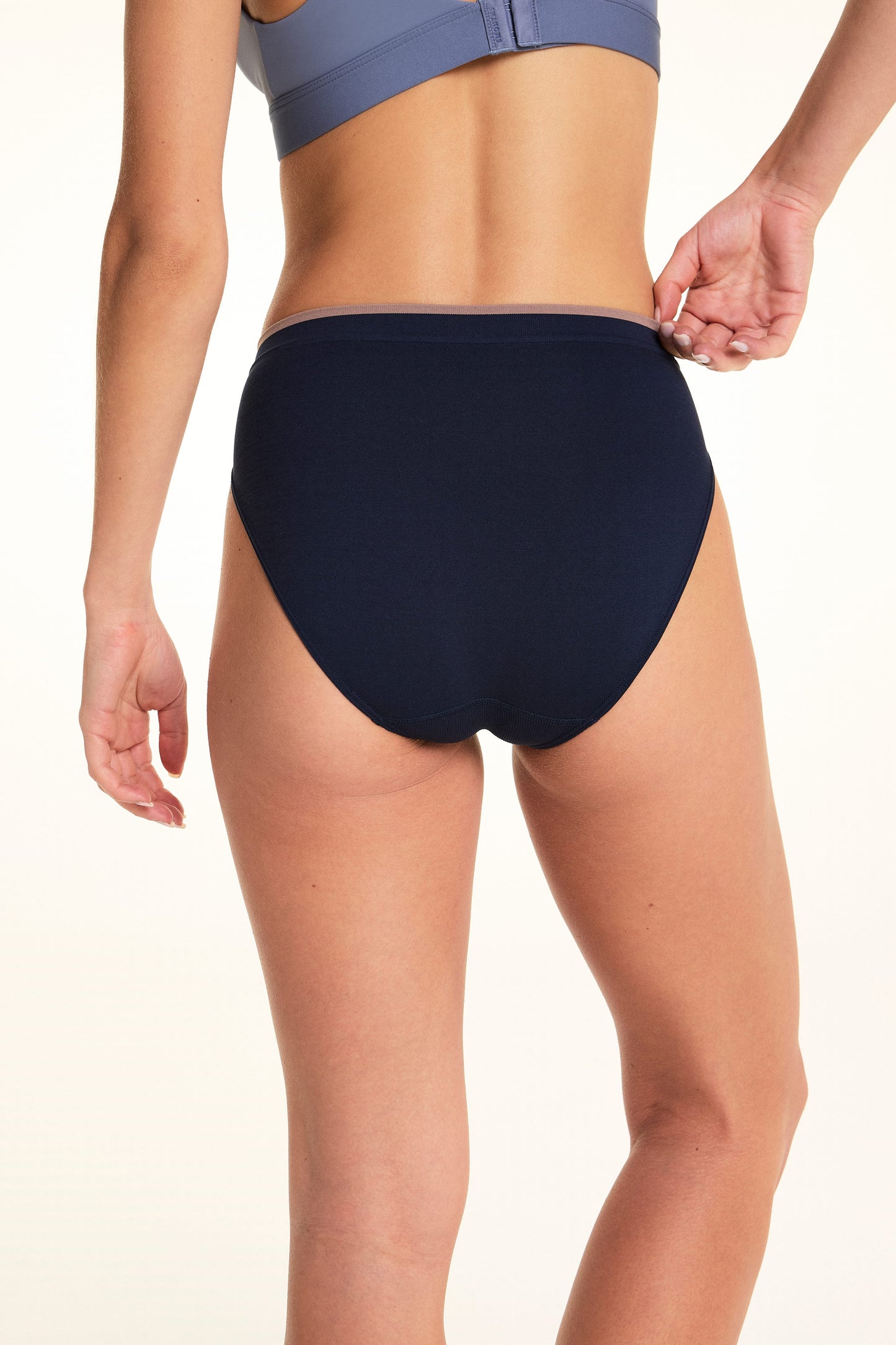 On a light studio backdrop, Alexis is shown from the back wearing the Seamless Brief in Navy. She gently pulls the waistband to demonstrate the smooth seamless fabric and full back coverage.