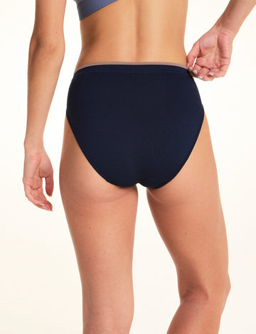 On a light studio backdrop, Alexis is shown from the back wearing the Seamless Brief in Navy. She gently pulls the waistband to demonstrate the smooth seamless fabric and full back coverage.