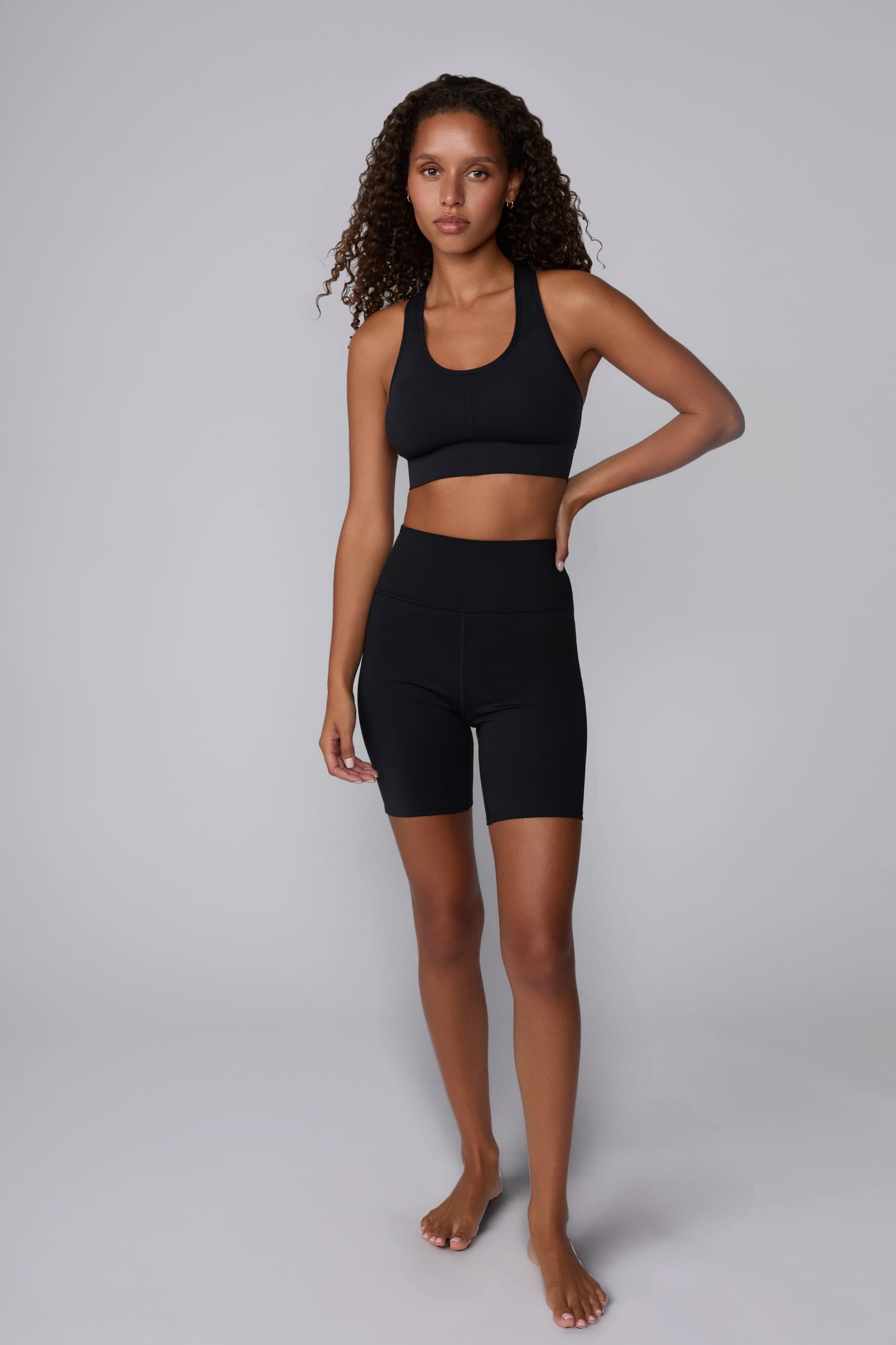 On a light gray studio backdrop, Courtney wears the Seamless Racerback Sports Bra in Black with matching bike shorts. The full-body view showcases the scoop neckline, racerback silhouette, wide underband, and seamless design paired with high-rise bike shorts.