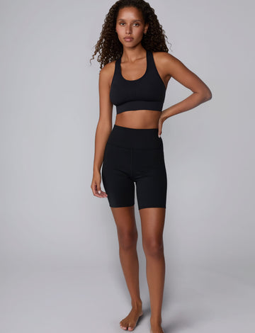 On a light gray studio backdrop, Courtney wears the Seamless Racerback Sports Bra in Black with matching bike shorts. The full-body view showcases the scoop neckline, racerback silhouette, wide underband, and seamless design paired with high-rise bike shorts.