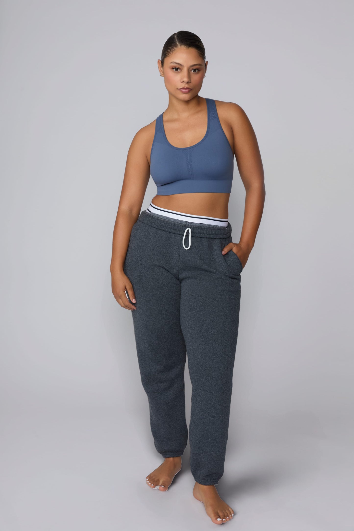 On a light gray studio backdrop, India wears the Seamless Racerback Sports Bra in Evernight with charcoal joggers. The full-body view showcases the scoop neckline, racerback silhouette, wide underband, and seamless design.