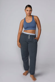 On a light gray studio backdrop, India wears the Seamless Racerback Sports Bra in Evernight with charcoal joggers. The full-body view showcases the scoop neckline, racerback silhouette, wide underband, and seamless design.