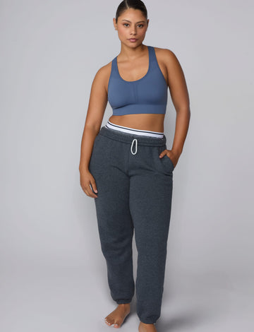 On a light gray studio backdrop, India wears the Seamless Racerback Sports Bra in Evernight with charcoal joggers. The full-body view showcases the scoop neckline, racerback silhouette, wide underband, and seamless design.