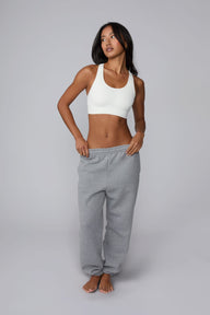 On a light gray studio backdrop, Amber wears the Seamless Racerback Sports Bra in White with gray sweatpants. The full-body view showcases the scoop neckline, racerback silhouette, wide underband, and seamless design.