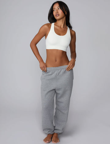On a light gray studio backdrop, Amber wears the Seamless Racerback Sports Bra in White with gray sweatpants. The full-body view showcases the scoop neckline, racerback silhouette, wide underband, and seamless design.