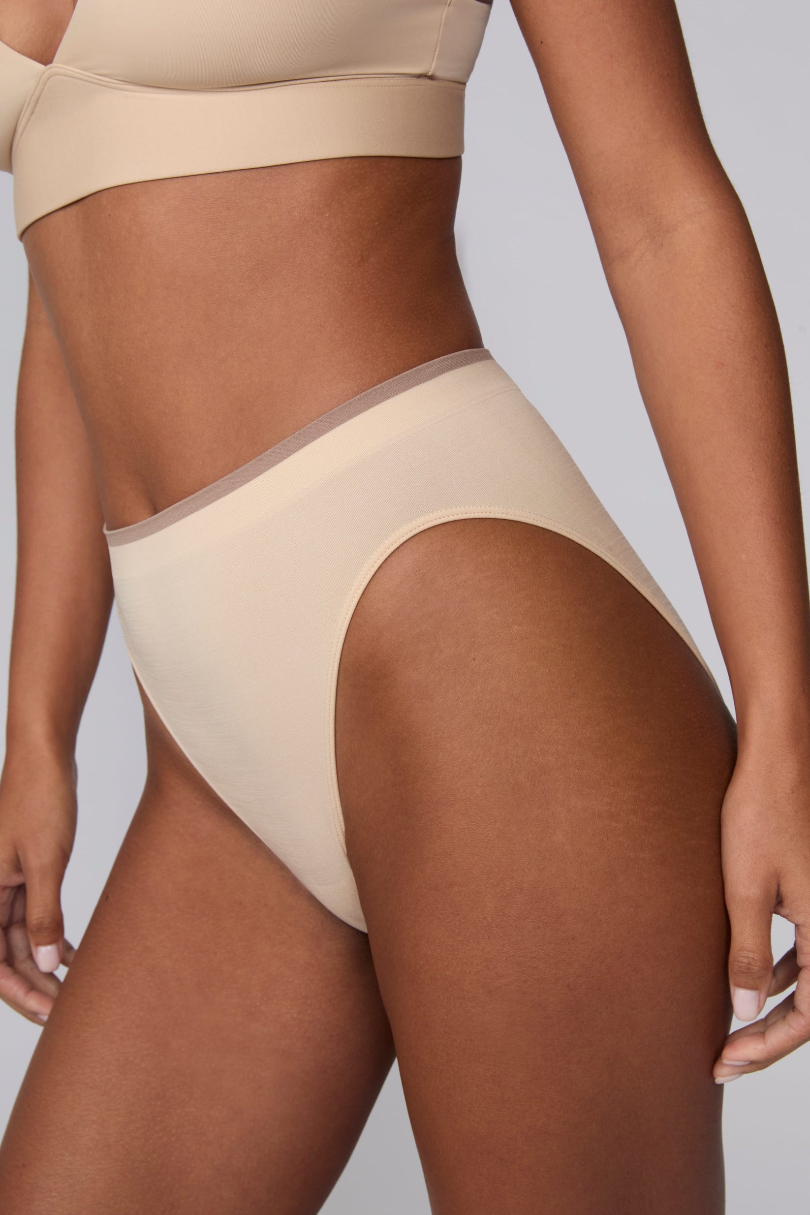 In a studio setting, Courtney wears the Smooth Seamless Brief in Beige. A close-up angled view highlights the high-leg cut, smooth fabric, and contrast trim waistband.