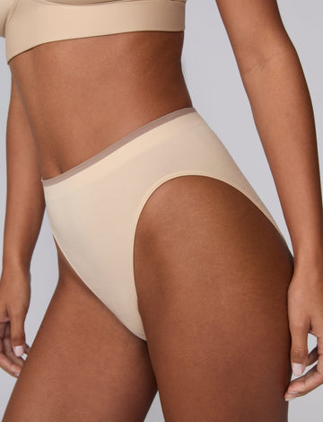 In a studio setting, Courtney wears the Smooth Seamless Brief in Beige. A close-up angled view highlights the high-leg cut, smooth fabric, and contrast trim waistband.