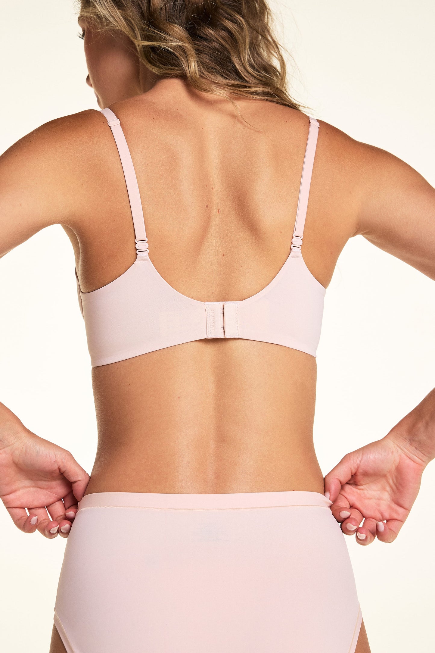 On a light neutral backdrop, Alexis is shown from the back wearing the Base Balconette Bra in Rose. The image highlights the adjustable straps, wide back band, and hook-and-eye closure.

