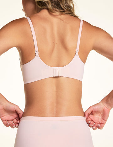 On a light neutral backdrop, Alexis is shown from the back wearing the Base Balconette Bra in Rose. The image highlights the adjustable straps, wide back band, and hook-and-eye closure.
