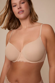 On a neutral studio backdrop, Sonnie wears the Base Lace T-Shirt Bra in Beige with matching underwear. The close-up view highlights the smooth molded cups, underwire support, adjustable straps, and delicate lace detail at the center front.