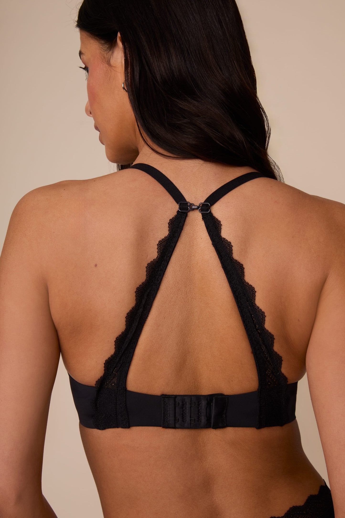 On a neutral studio backdrop, Marcela wears the Base Lace T-Shirt Bra in Black. The back view highlights the convertible racerback feature, adjustable straps, hook-and-eye closure, and lace detailing along the racerback panel.
