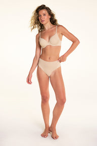 On a light studio backdrop, Alexis wears the Base Picot T-Shirt Bra in Beige with matching beige underwear. The full-body image highlights the smooth molded cups, picot trim along the neckline, and adjustable straps.