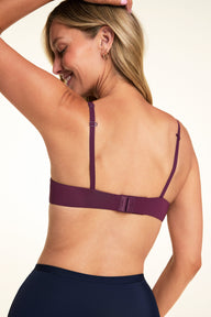 On a light studio backdrop, Meagan wears the Base T-Shirt Bra in Eggplant. The angled back view showcases the smooth band, adjustable straps, and hook-and-eye closure.
